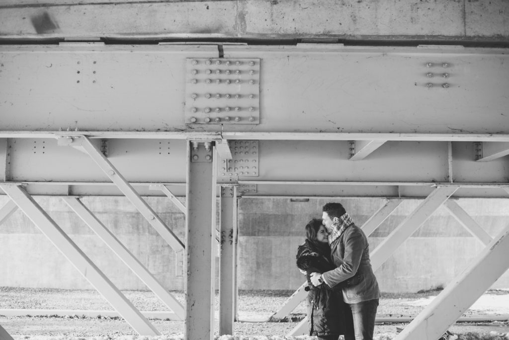 Black and white photo of a couple in winter coats embracing beneath the steel beams of the bridge at Bate Island in Ottawa.