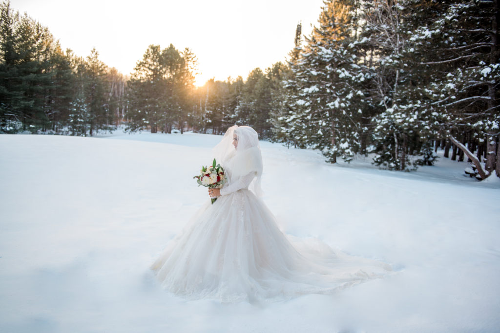 Bride in a full lace gown and veil standing in deep snow at Pinhey Forest on the NCC Trails in Nepean, holding a red and white bouquet as the sun sets behind snow-covered pines.