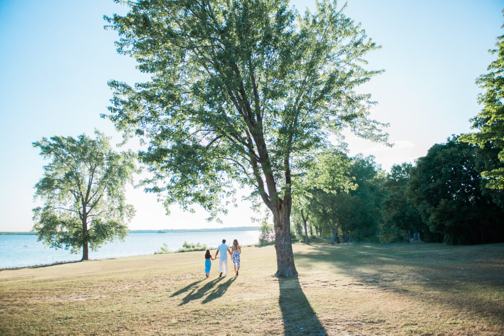 Family of three holding hands and walking toward the water under a large tree at Aylmer Marina in Quebec, with long shadows across the grass in the summer light.