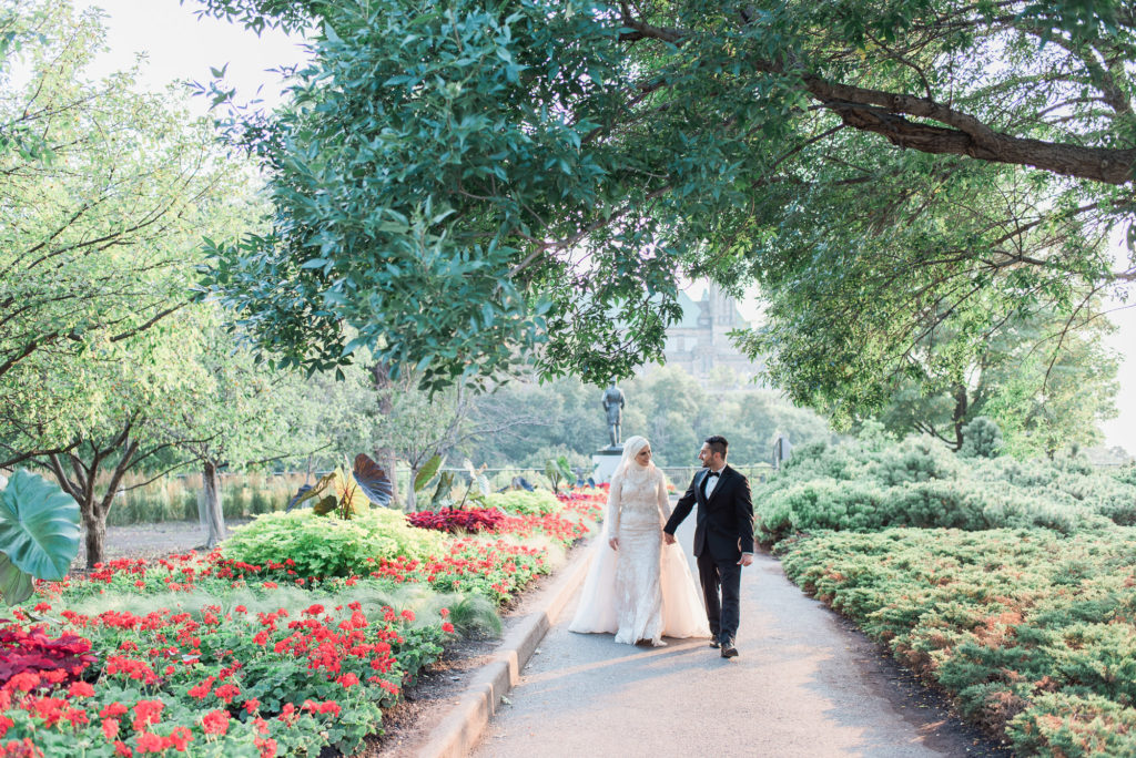 Bride and groom holding hands as they walk a garden path lined with red flowers at Major's Hill Park in Ottawa, the bride in a lace gown and hijab and the groom in a black tuxedo.