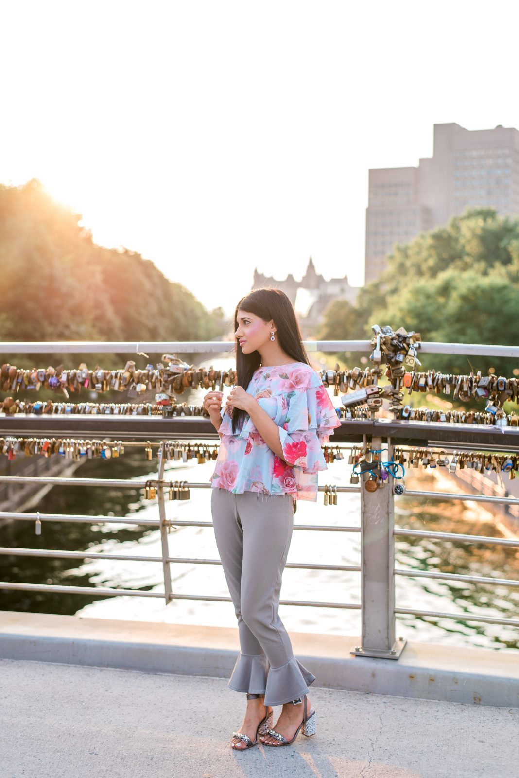 Woman in a floral top and grey ruffled pants standing on the love lock bridge over the Rideau Canal at the University of Ottawa campus, backlit by golden sunset light.