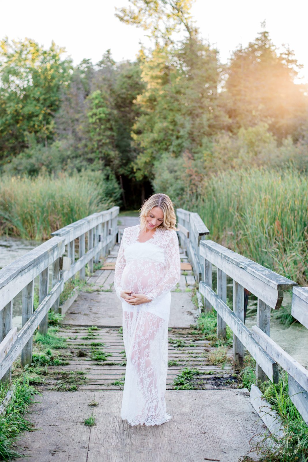 Expecting mother in a white lace maternity gown standing on a wooden boardwalk surrounded by tall reeds at Baxter Conservation Area near Ottawa, backlit by golden sunset light.