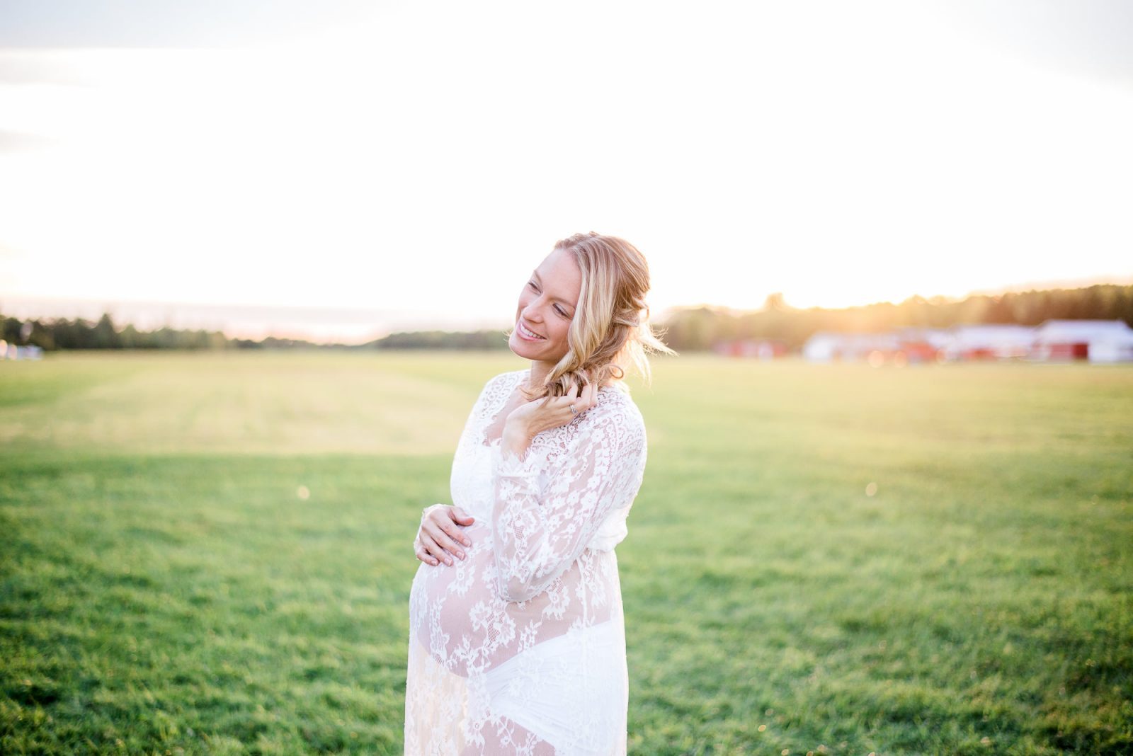 Expecting mother in a white lace gown smiling and cradling her bump in an open field at Baxter Conservation Area near Ottawa, with warm sunset light behind her.