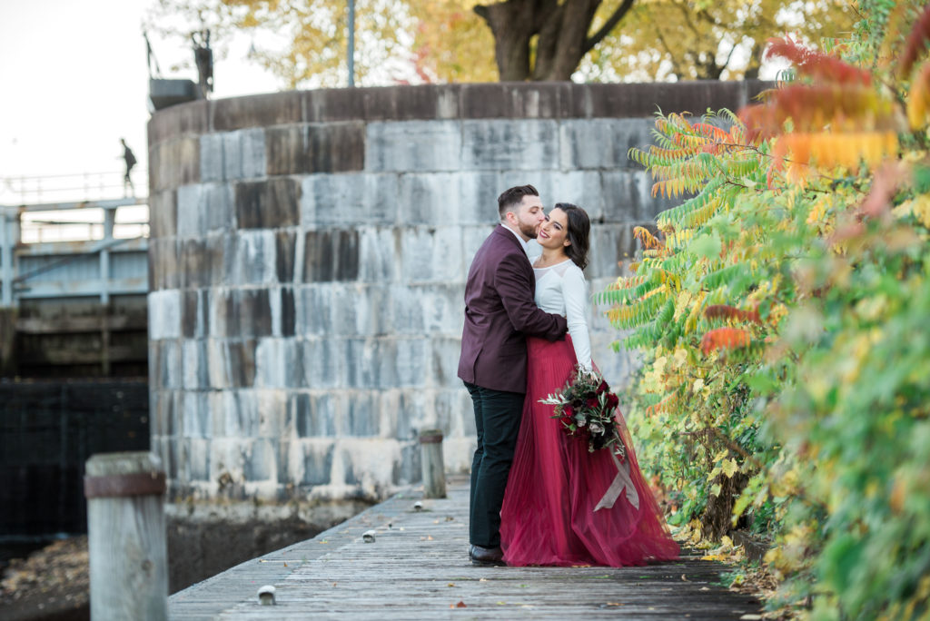 Couple embracing on a wooden walkway beside the stone lock walls at Hog's Back Park in Ottawa, the woman in a deep red tulle skirt holding a bouquet, surrounded by fall foliage.