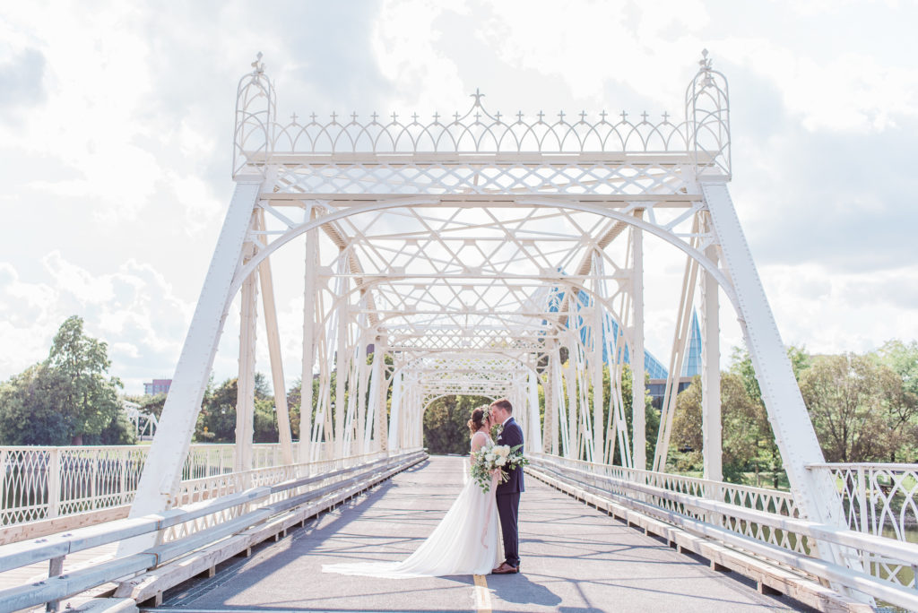 Bride and groom standing forehead to forehead in the centre of the white iron Minto Bridges near Stanley Park in Ottawa, framed by the bridge's lattice arches under a cloudy blue sky.