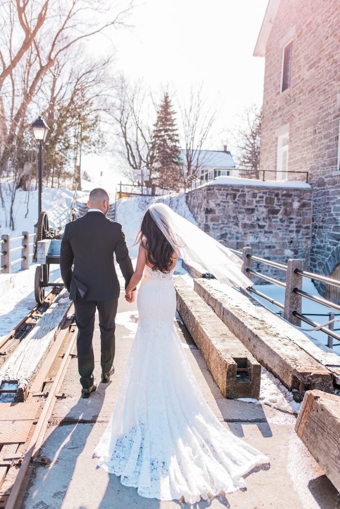 Bride and groom walking hand in hand past the stone facade of Watson's Mill in Manotick, Ottawa, snow along the riverbank and her veil catching the wind.
