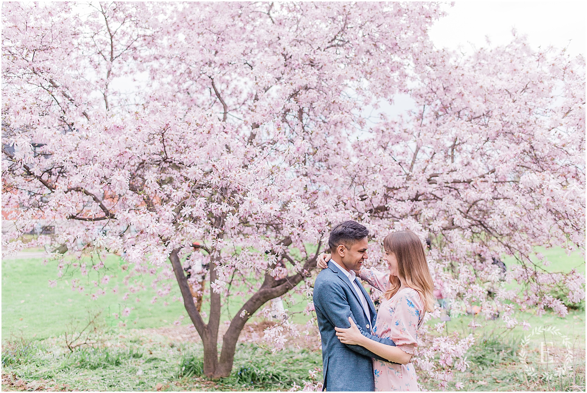 Couple embracing and smiling at each other beneath a blossoming pink tree at the Ornamental Gardens of the Central Experimental Farm in Ottawa, the man in a blue blazer and the woman in a soft pink floral dress.