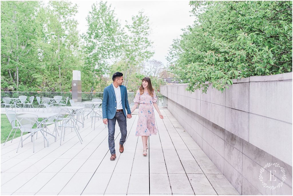 Couple walking hand in hand along the outdoor terrace at the Canadian Museum of Nature in Ottawa, the man in a blue blazer and the woman in a pink floral dress, with white patio chairs and stone walls beside them.