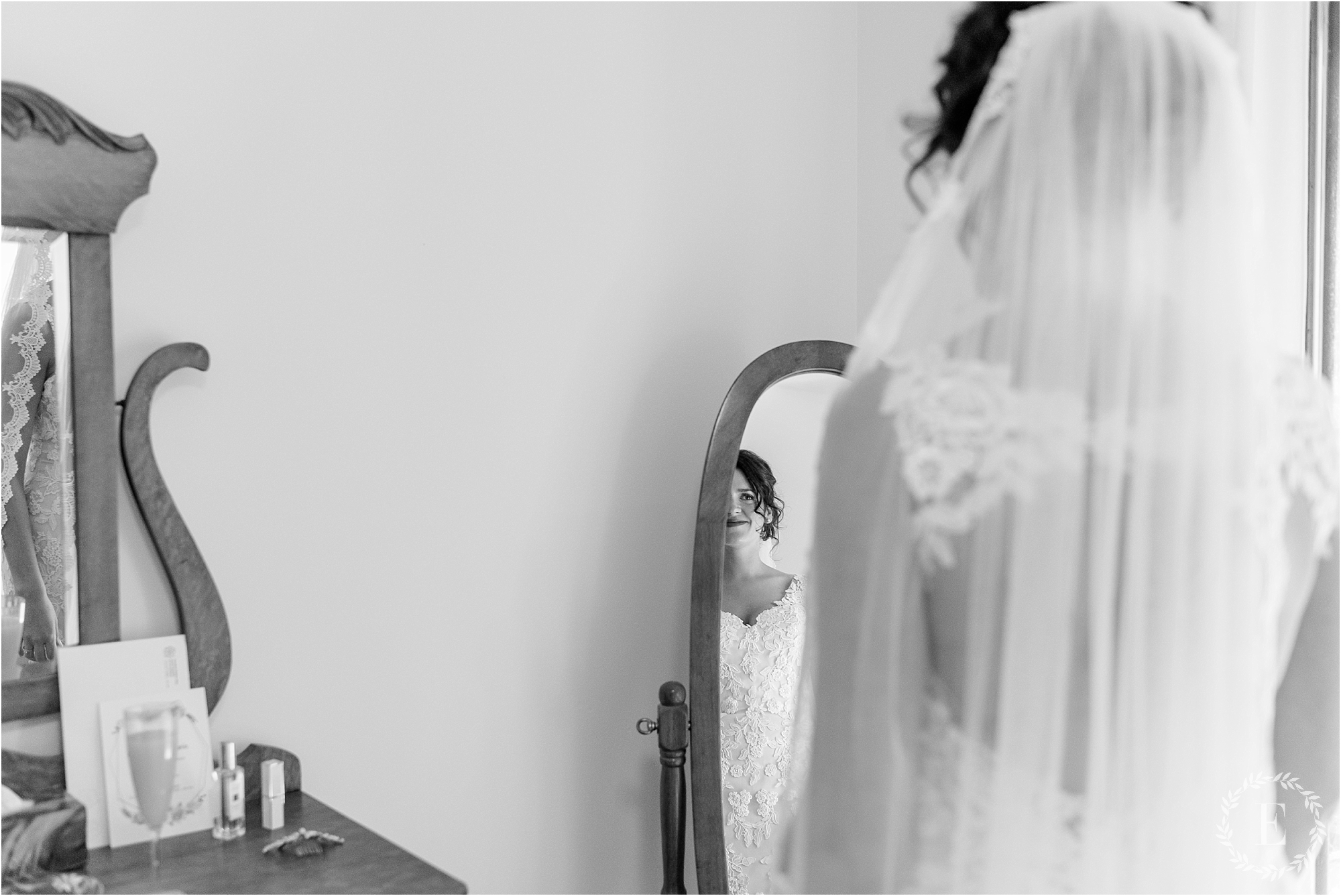Black and white photo from Revelle Bridal showing a bride's reflection in an antique wooden mirror, wearing a long lace veil, with a vanity table visible below