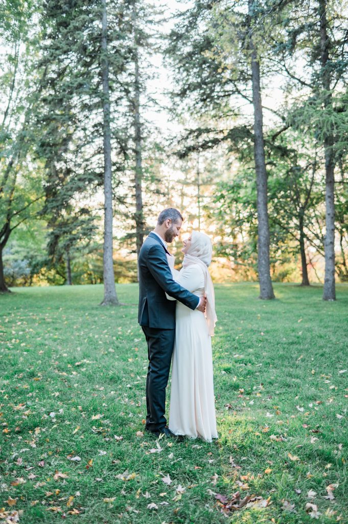 Couple standing forehead to forehead among tall pines at Claudette Cain Park in Ottawa, the woman in a cream gown and hijab and the man in a navy suit, with fallen autumn leaves on the grass.