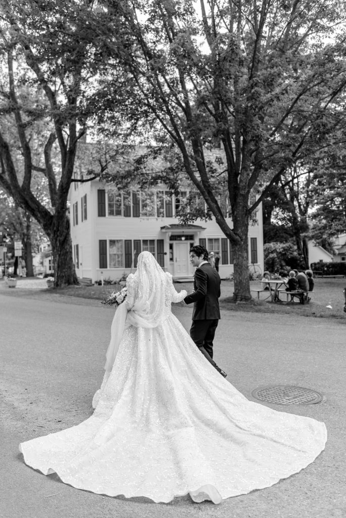 Black and white photo of a bride and groom walking together in front of the Dickinson House in Manotick, Ottawa, her long beaded train trailing behind her on the road.