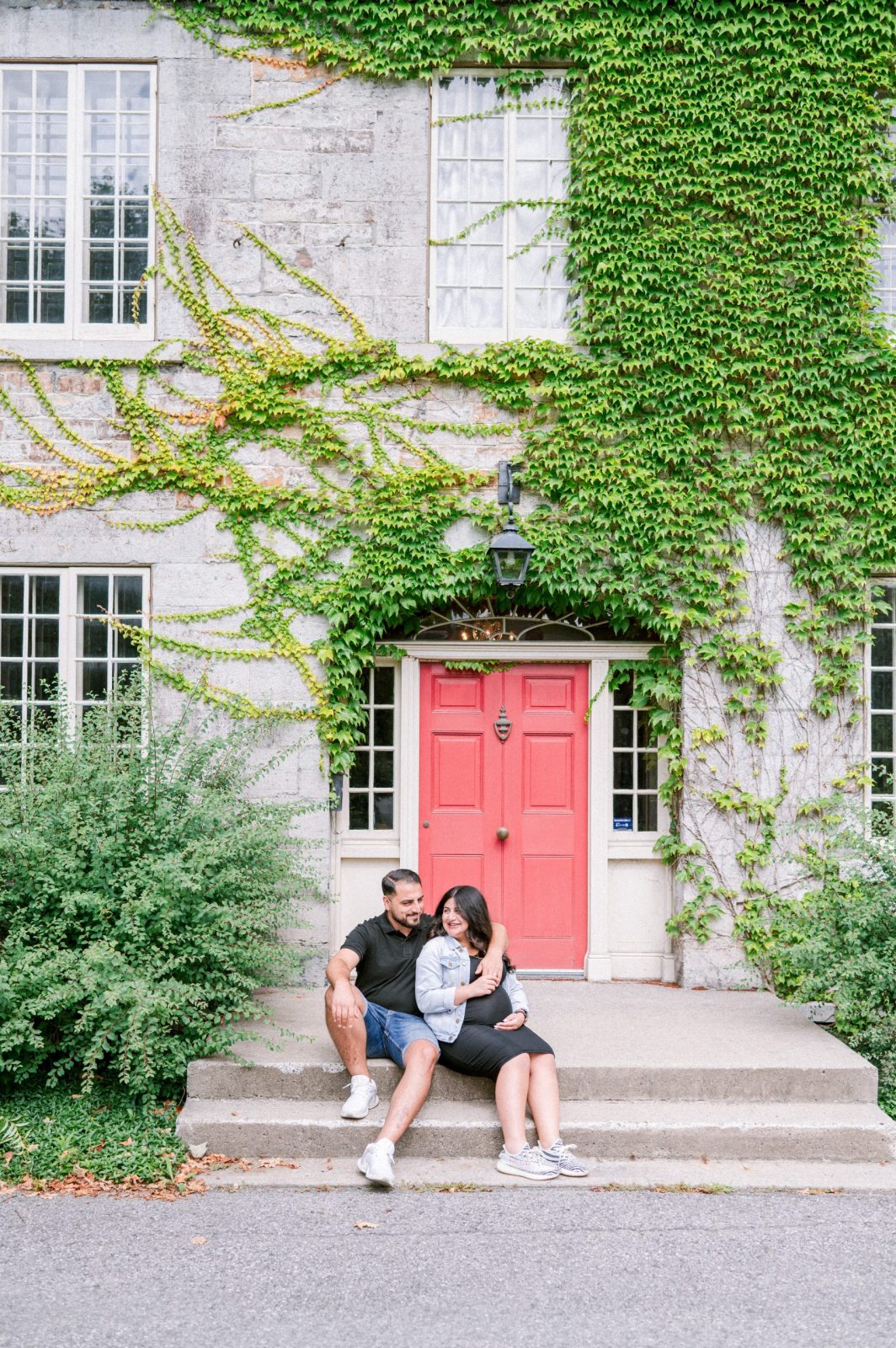 Expecting couple sitting on the front steps of the ivy-covered stone Keg Manor in Ottawa, beneath its bright pink door.
