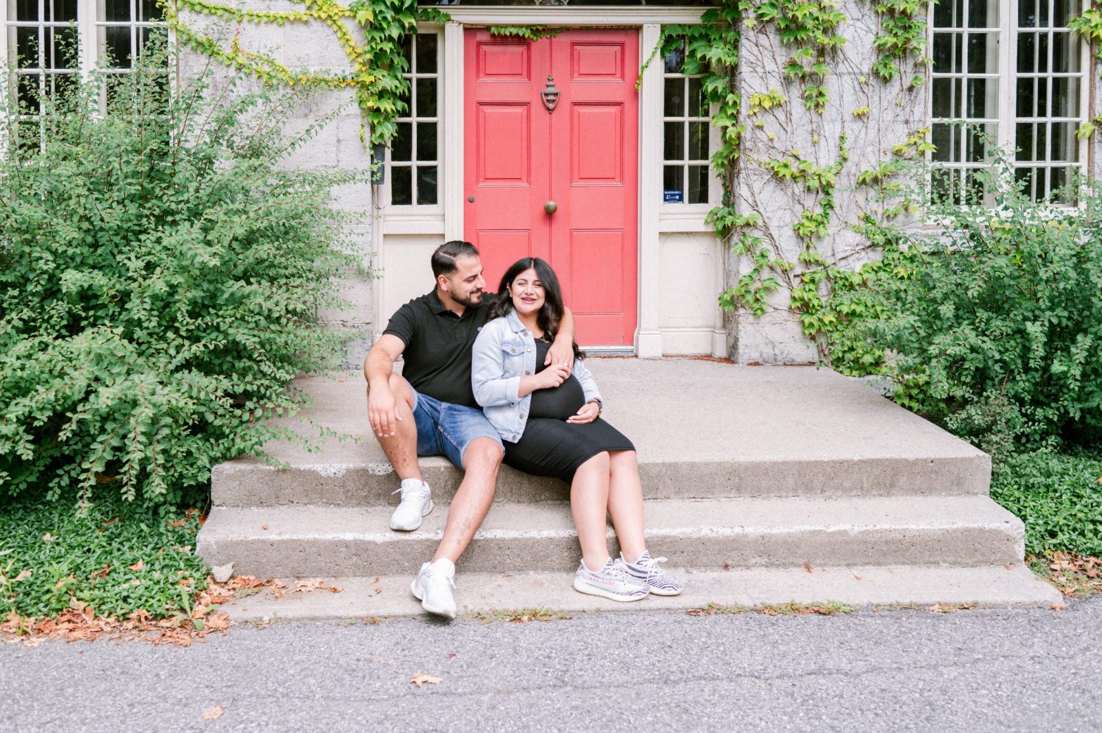 Close-up of an expecting couple sitting together on the steps of the Keg Manor in Ottawa, with the pink door and climbing ivy behind them and his hand resting on her bump.