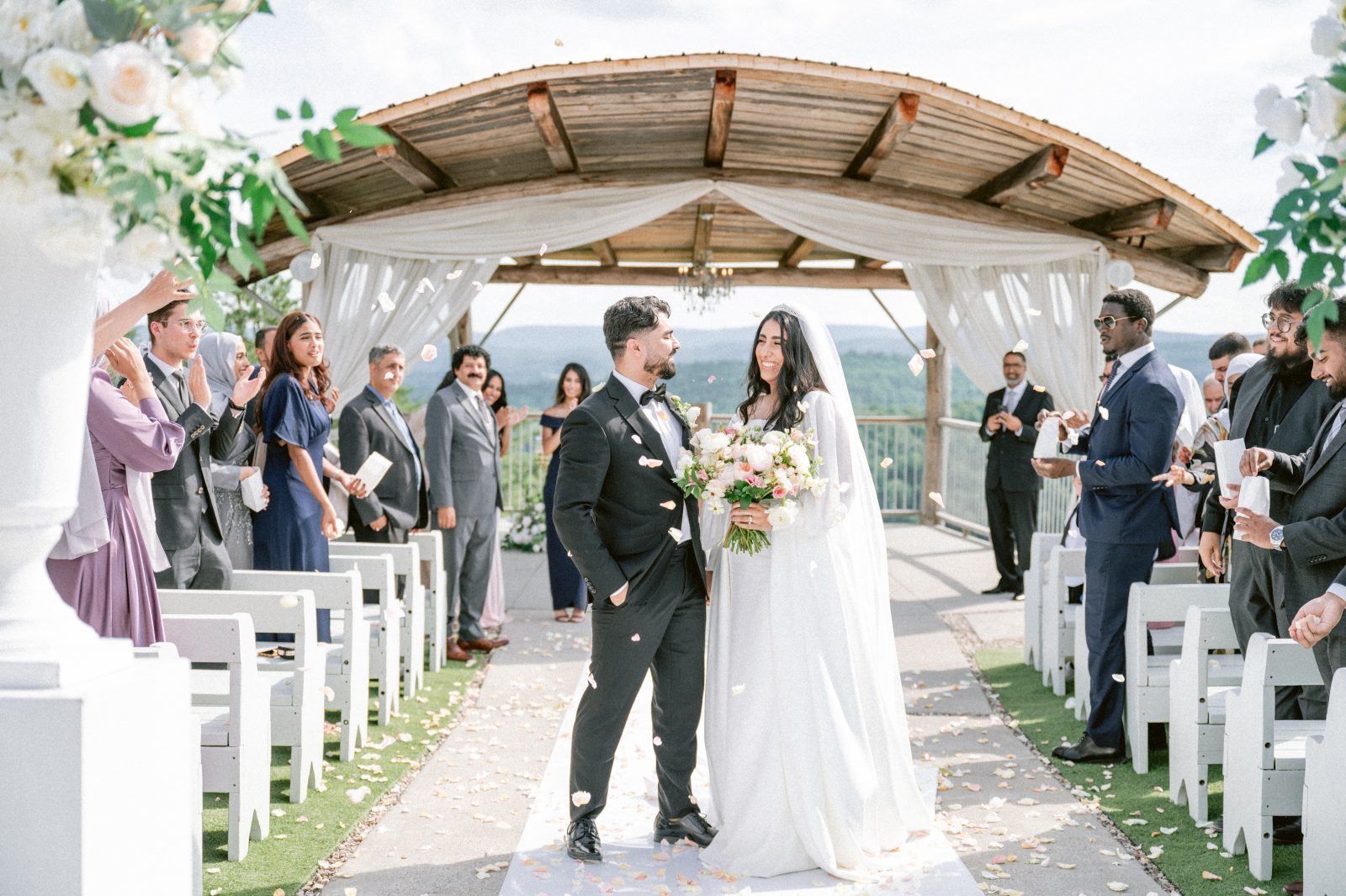 Newlyweds walking down the aisle under a wooden pergola as guests throw flower petals, with a scenic overlook of hills in the background.