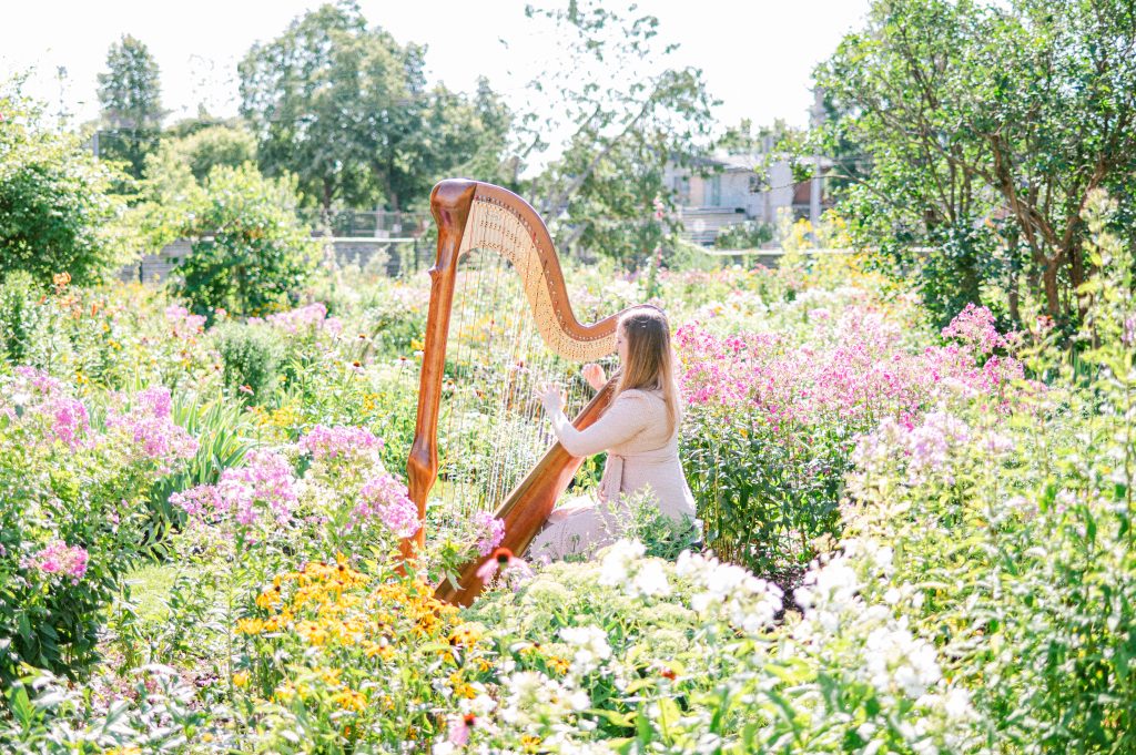 Woman in a cream outfit playing a wooden harp while surrounded by tall pink and yellow blooms at Maplelawn Garden in Ottawa.