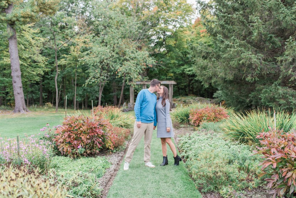 Couple kissing in the gardens at the Mackenzie King Estate in Gatineau Park, surrounded by autumn shrubs and tall trees, with a stone ruin behind them.