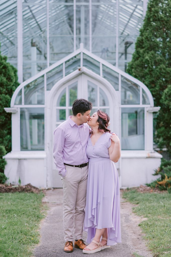 Couple in lavender kissing in front of the arched white Tropical Greenhouses at the Central Experimental Farm in Ottawa.