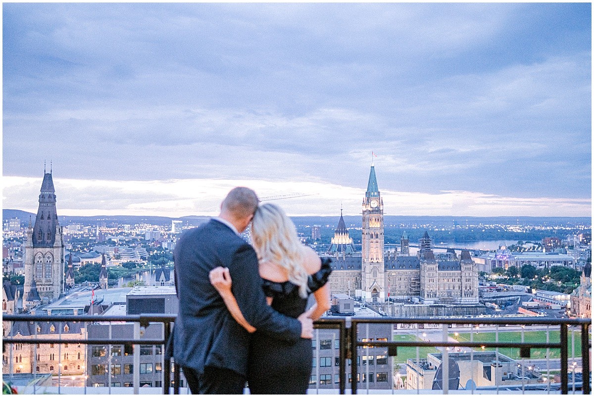 Couple embracing on a rooftop terrace overlooking the city skyline at dusk, with illuminated Gothic towers in the distance.