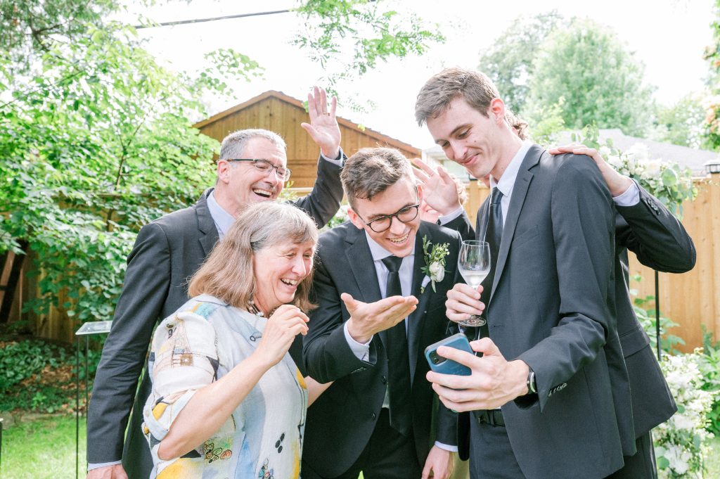 Guests and groomsmen laughing together while looking at a phone during an outdoor backyard wedding celebration.
