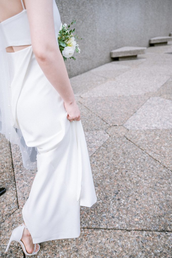 Close-up of a bride from White Satin Bridal in a minimalist satin gown with a cutout back, holding the fabric while walking on stone pavement, a small bouquet visible on her shoulder
