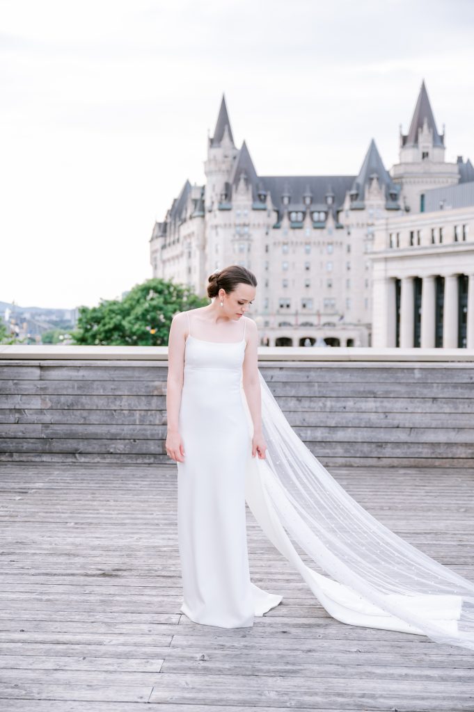 Bride from White Satin Bridal wearing a sleek square-neck satin gown with a long attached train, standing on a wooden boardwalk with a castle-like building in the background