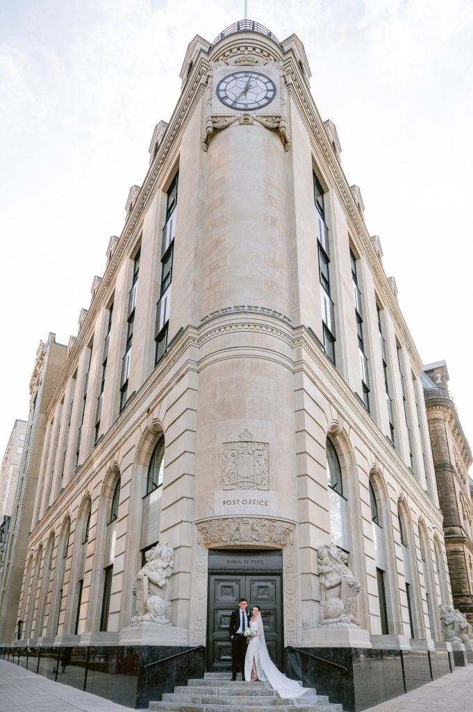 Bride and groom on the steps of the historic Canada Post building on Sparks Street, Ottawa, dwarfed by its clock tower and stone facade.
