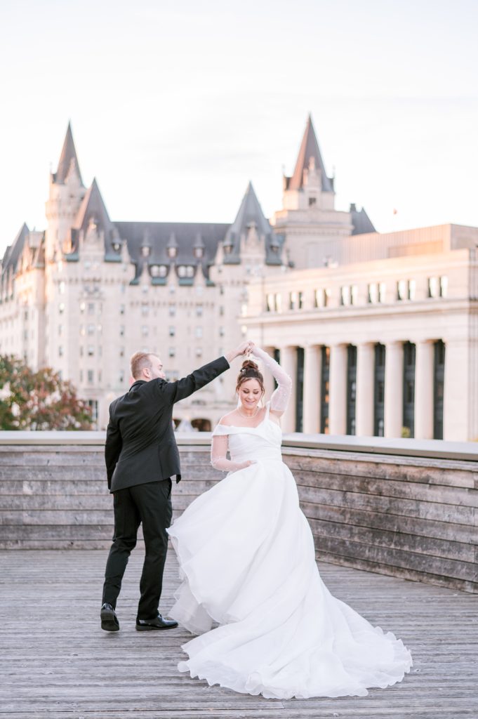 Groom twirling his bride on the wooden terrace at the National Arts Centre in Ottawa, with the Château Laurier rising behind them in warm evening light.