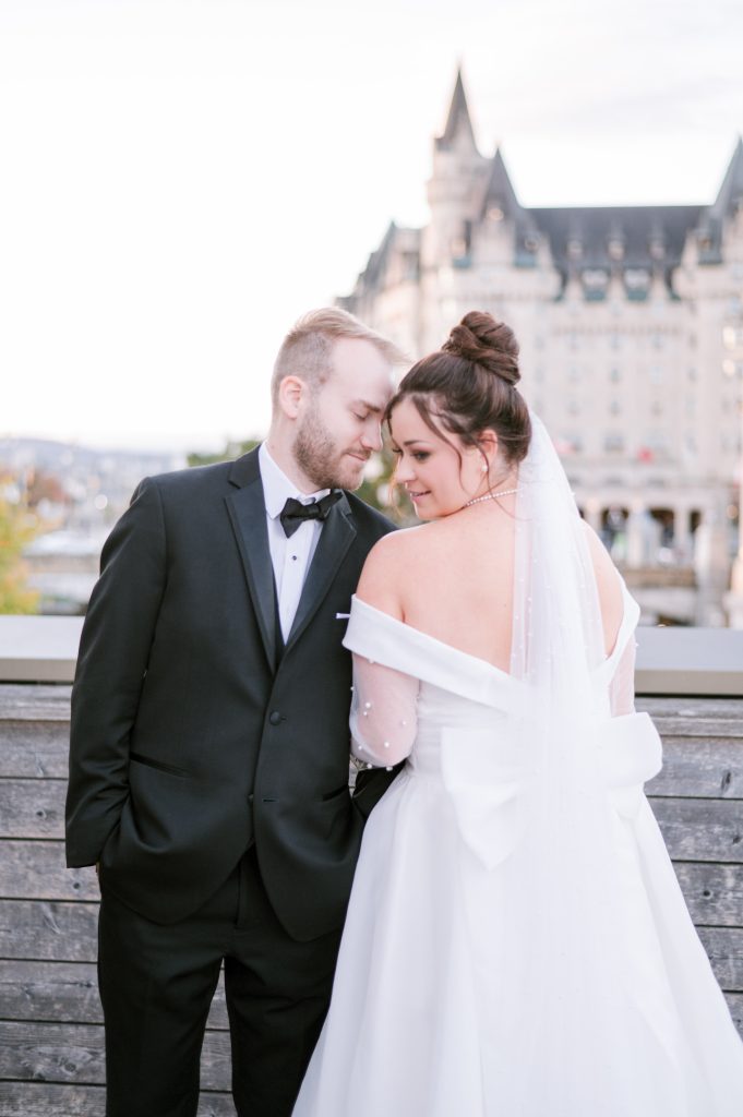 Bride and groom leaning close together on the terrace at the National Arts Centre in Ottawa, the Château Laurier visible behind them and her bow-back gown and veil in view.