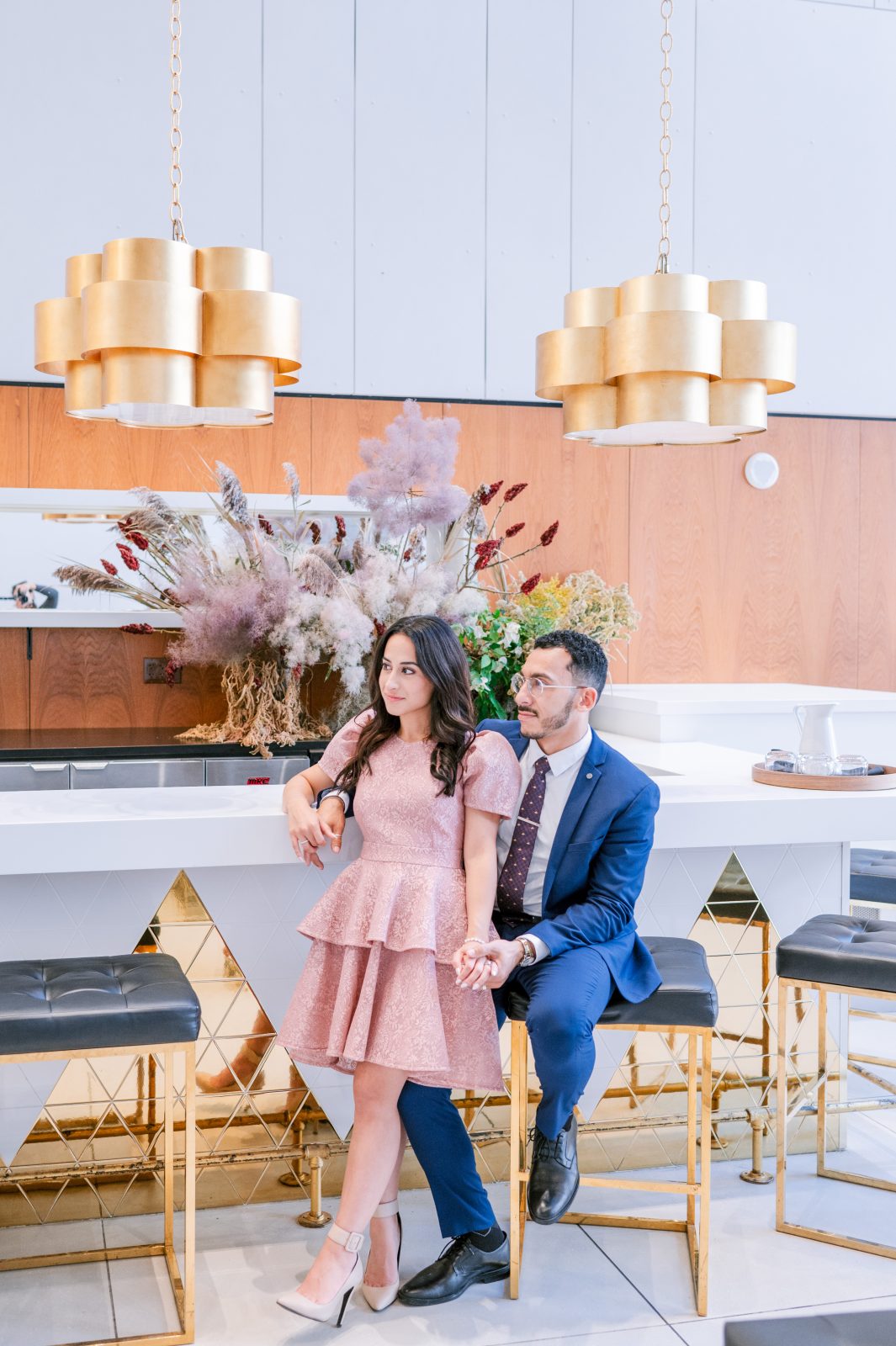 Couple posing at a modern bar area at the Ottawa Art Gallery, with gold pendant lights and a dried floral arrangement in the background.