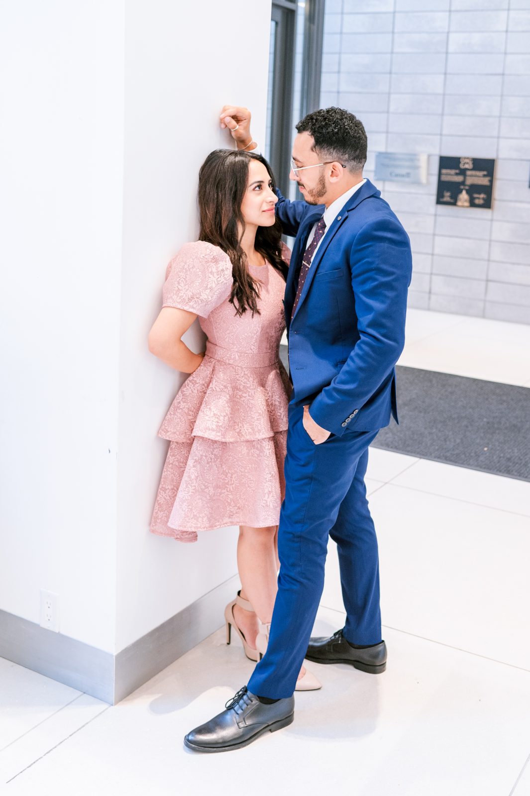Couple sharing a playful moment against a white wall at the Ottawa Art Gallery, the woman in a pink tiered dress and the man in a navy suit.