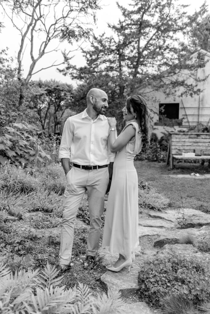 Black and white photo of a couple standing close together among the rocks and ferns at Fletcher Wildlife Garden in Ottawa, with an old barn in the background.