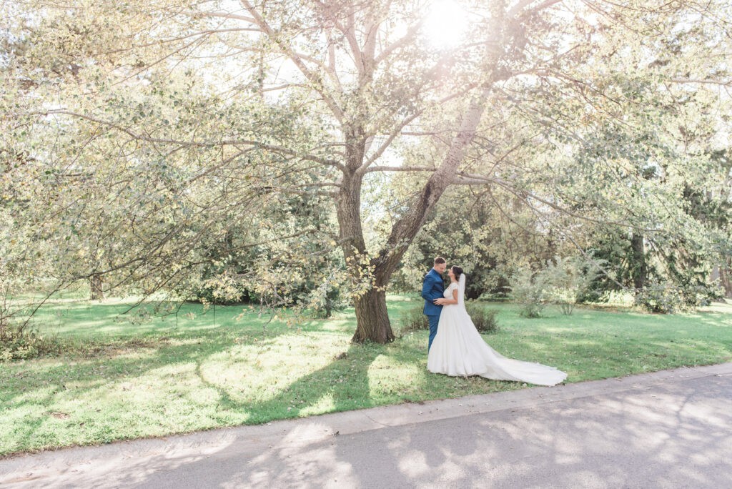 Bride and groom embracing beneath a large blossoming tree at the Dominion Arboretum near Dow's Lake in Ottawa, her long train spread across the grass in the afternoon sun.