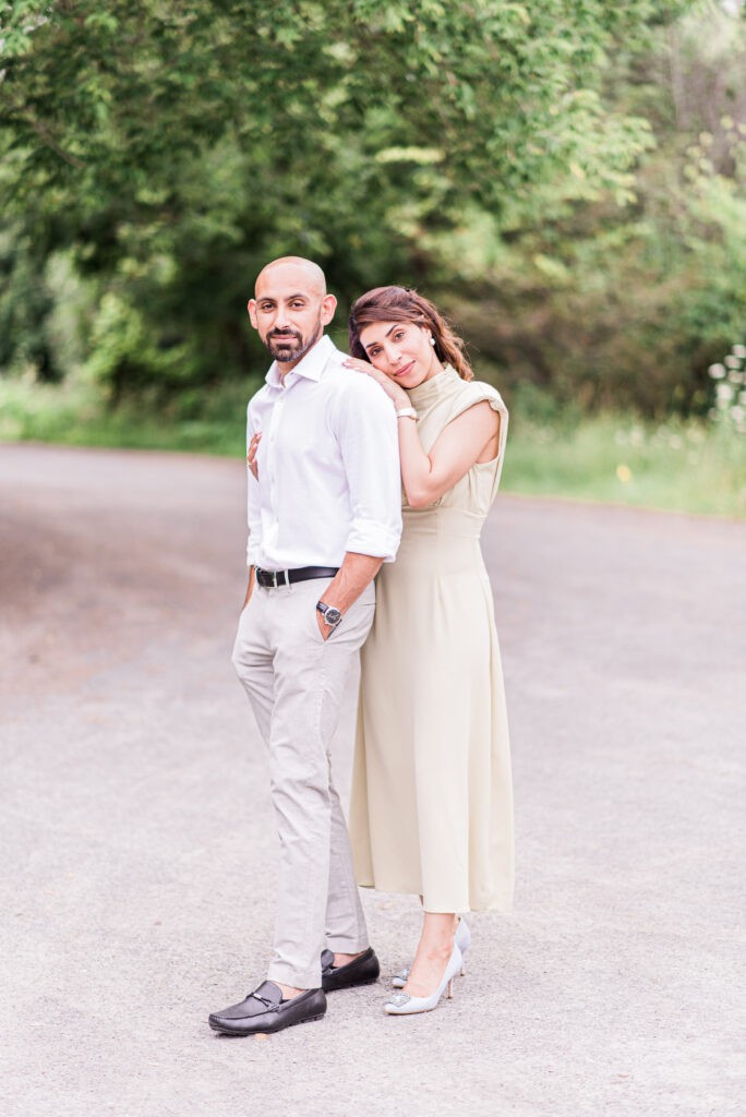 Couple posing on a gravel path at Fletcher Wildlife Garden in Ottawa, the woman in a sage green dress resting on the man's shoulder, with greenery behind them.