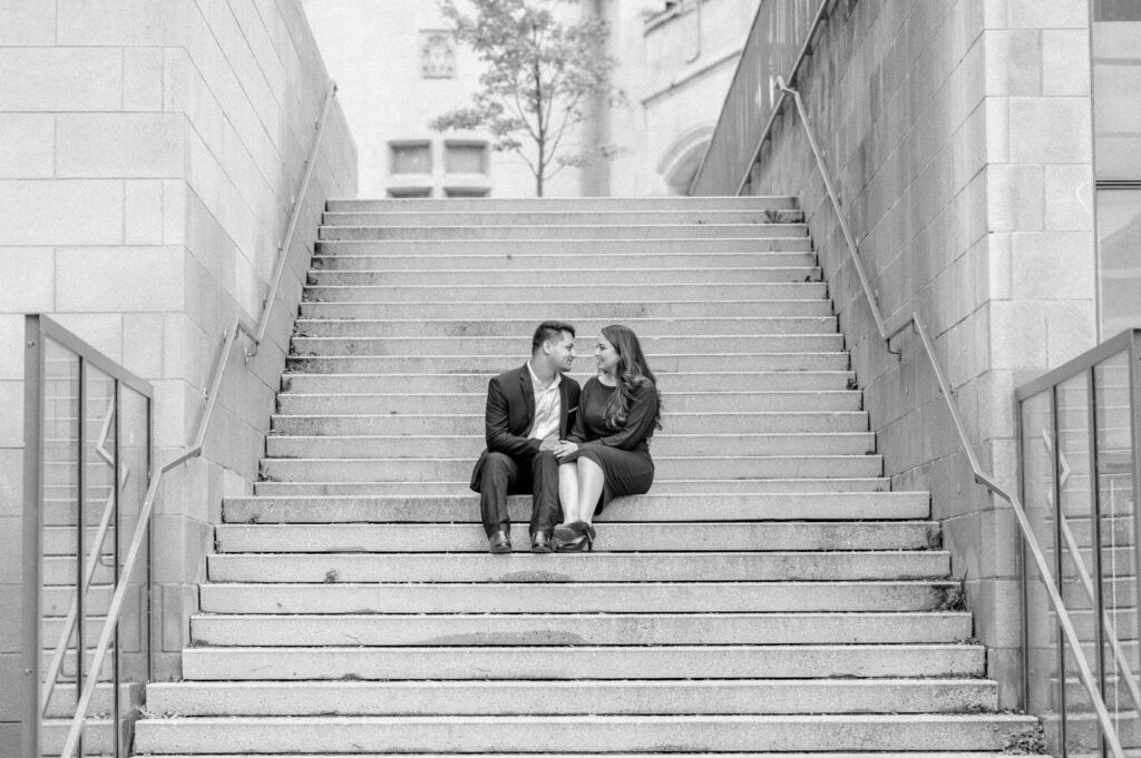 Black and white photo of a couple sitting close together on the York Steps in downtown Ottawa, framed by the long stone staircase and surrounding buildings.