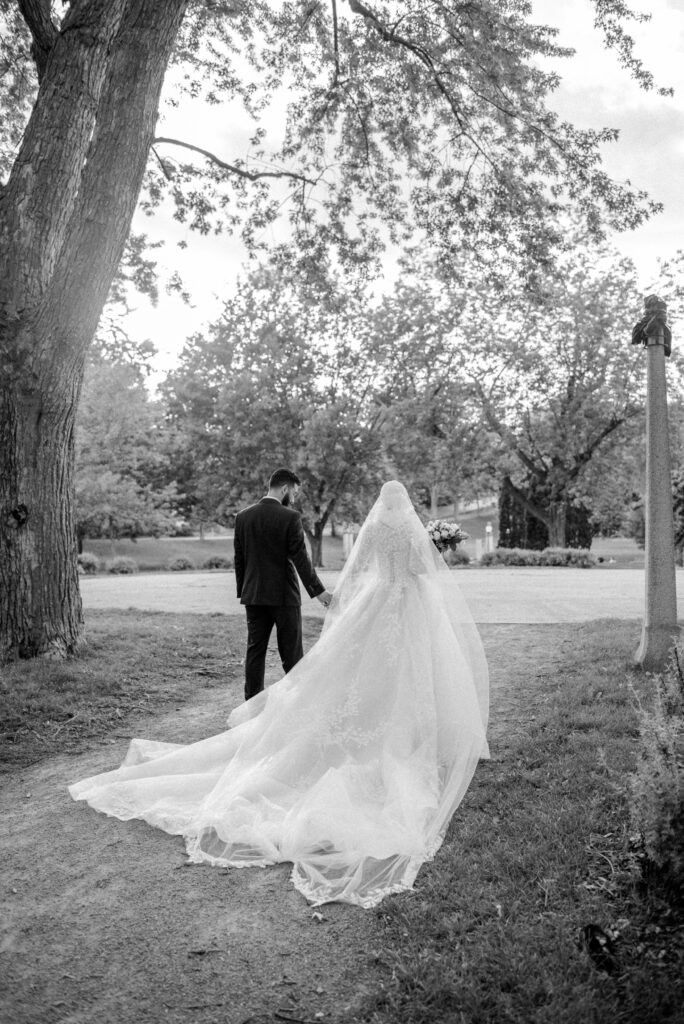 Black and white photo of a bride and groom walking away hand in hand under the trees at Strathcona Park in Ottawa, her long lace veil trailing across the grass behind her.