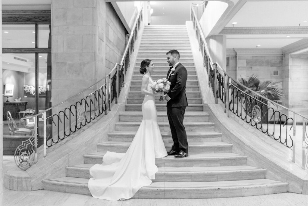 Black and white photo of a bride and groom standing together on a grand marble staircase in an Ottawa hotel lobby, her train cascading down the steps.