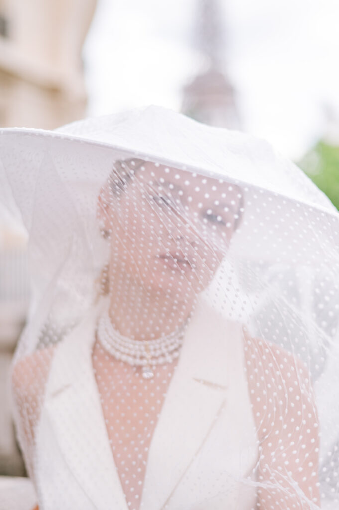 Woman in a wide-brimmed polka-dot veiled hat and pearl necklace poses with the Eiffel Tower faintly visible in the blurred background behind her.