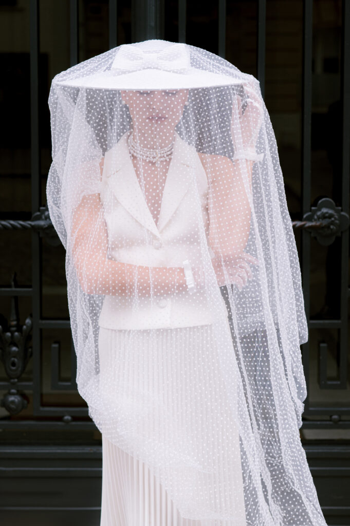 Woman in a white pleated skirt suit and gloves, arms crossed, wearing a wide-brimmed polka-dot veiled hat, standing before an ornate iron gate.