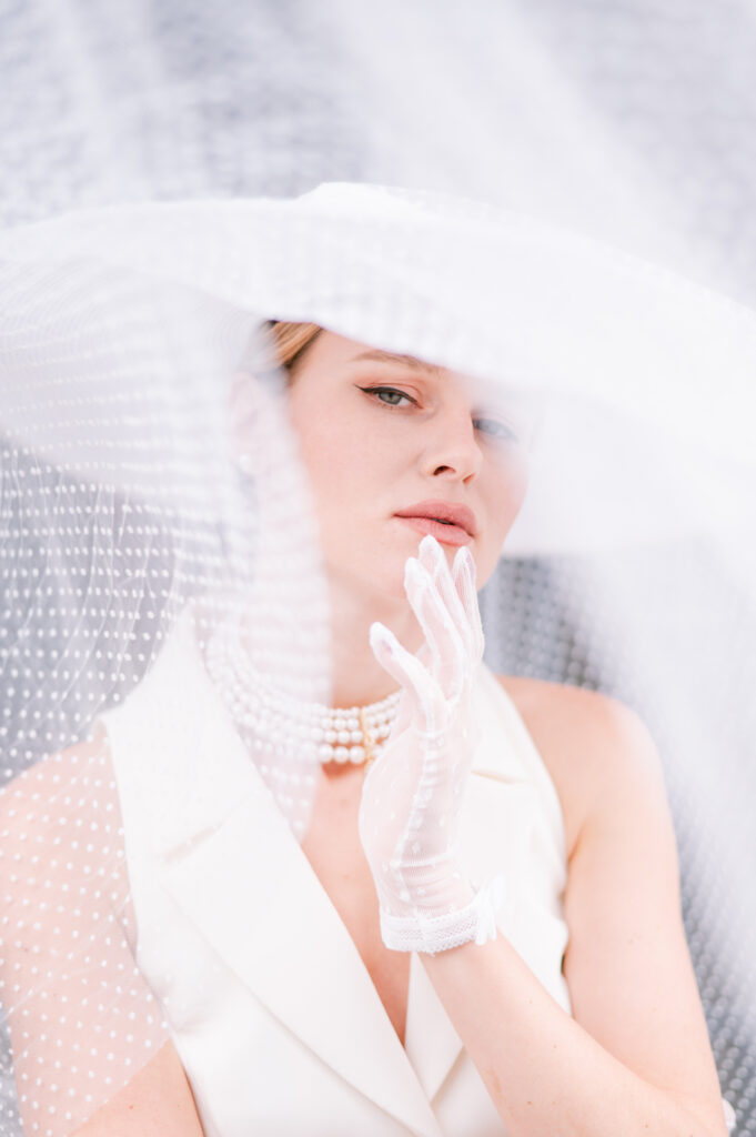 Woman in a wide-brimmed polka-dot veiled hat and white gloves touches her chin, wearing pearl chokers and a white blazer, with soft light filtering through the sheer veil.