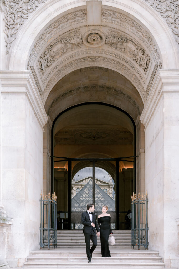 Couple in formal black attire walking through a grand stone archway entrance in Paris.