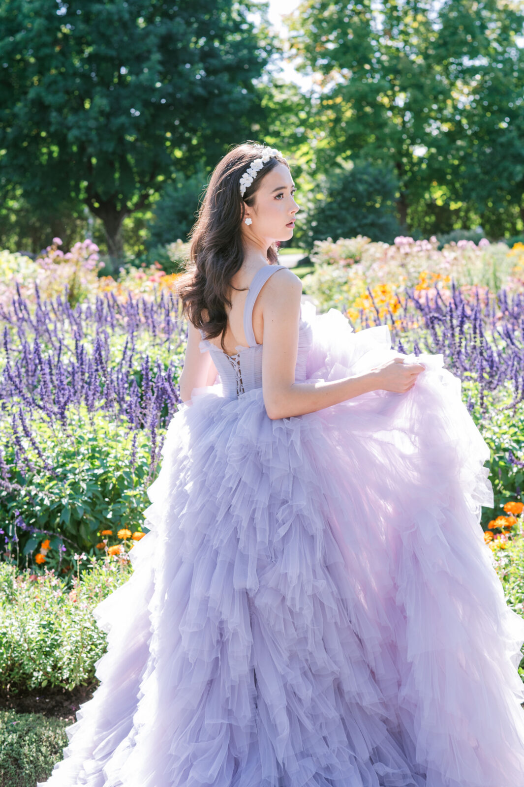 Model in a lavender tulle ball gown from Chic Bridals standing in a lush garden filled with purple and orange flowers, holding the voluminous skirt