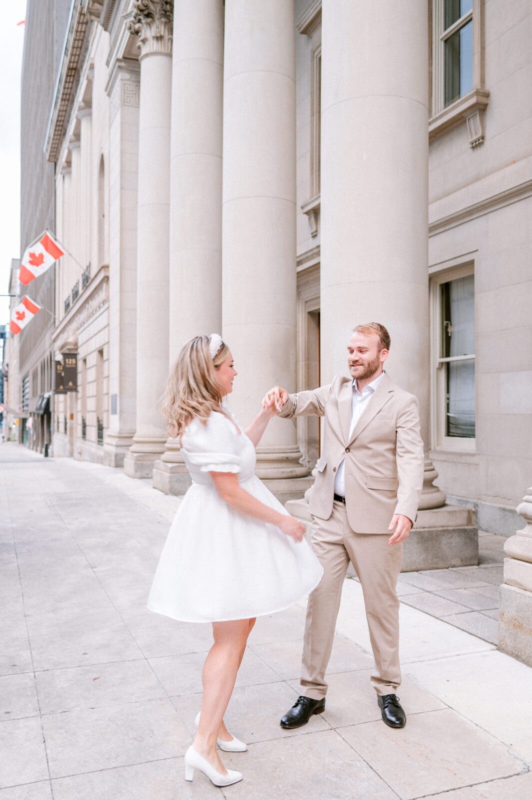 Couple dancing together on a city sidewalk near a Canadian flag and stone columned building.