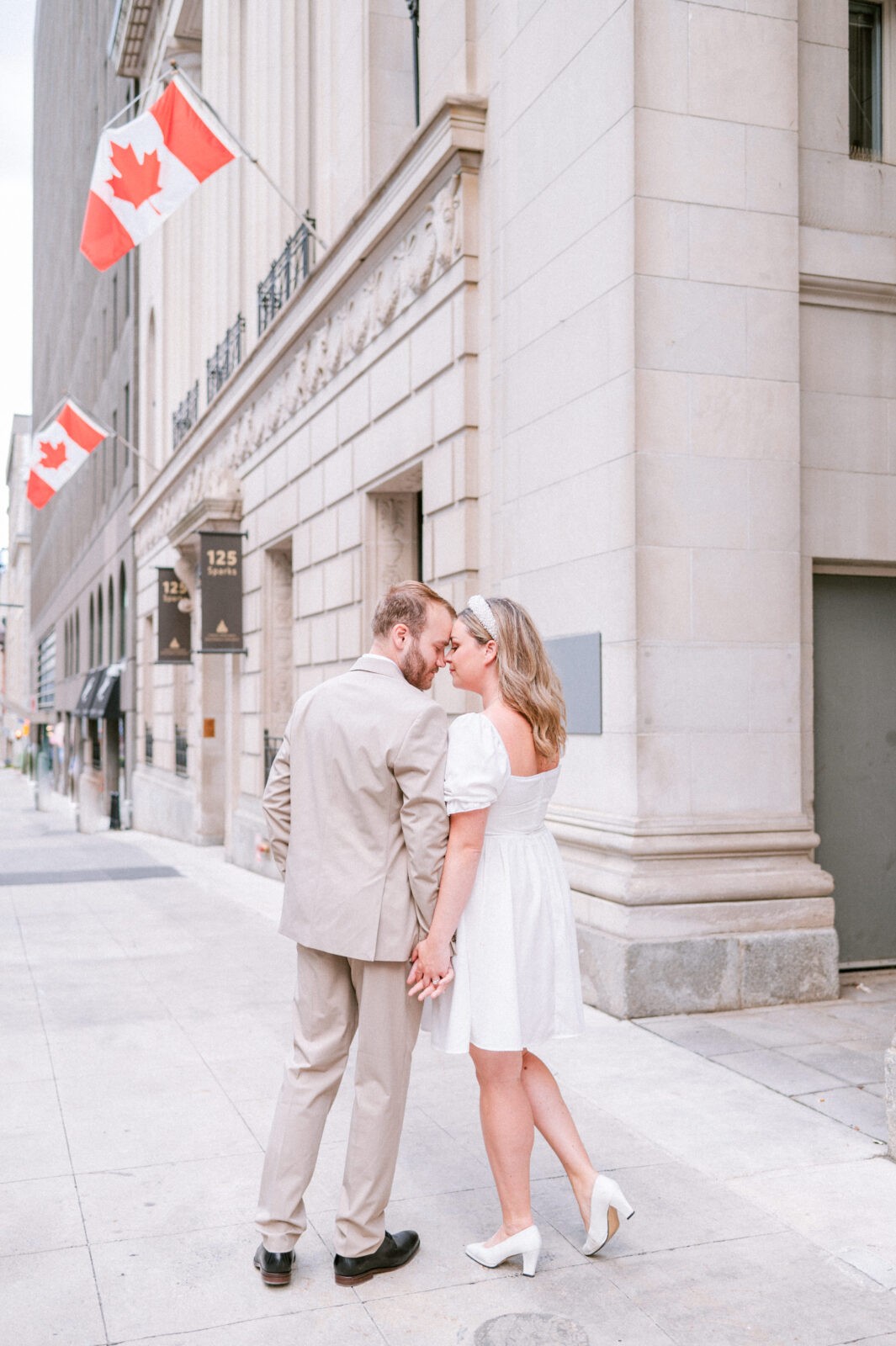 Couple sharing a close moment on a city sidewalk beneath Canadian flags and stone columned architecture.