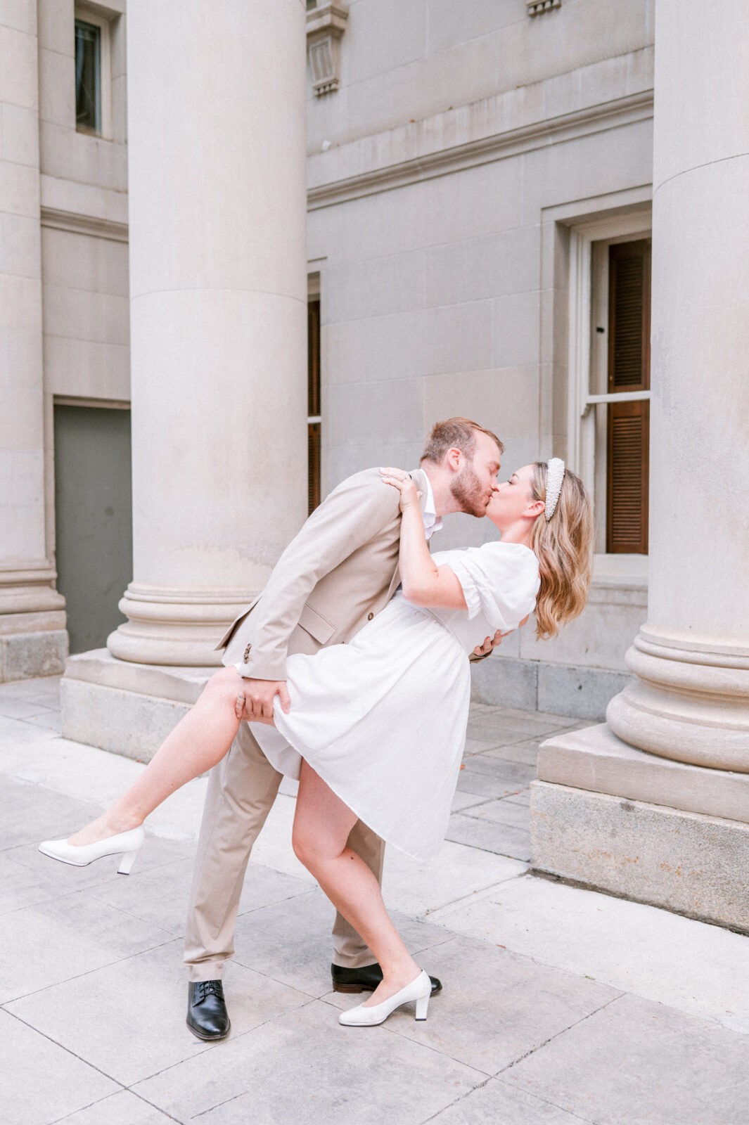 Groom dipping his partner for a playful kiss in front of a row of tall stone columns.