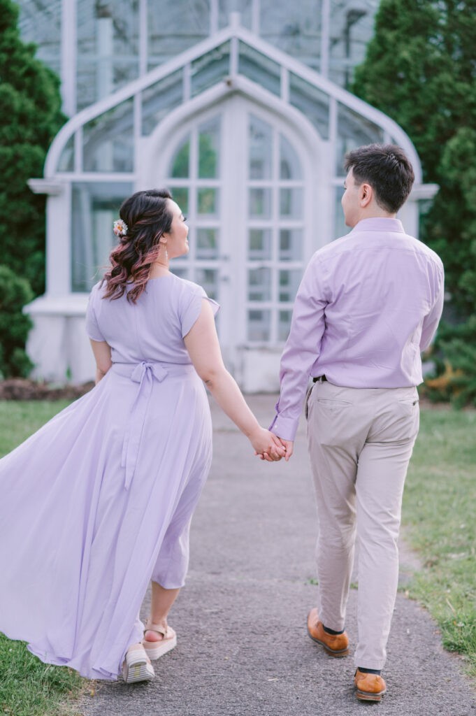 Couple in matching lavender outfits walking hand in hand toward the white glass Tropical Greenhouses at the Central Experimental Farm in Ottawa.