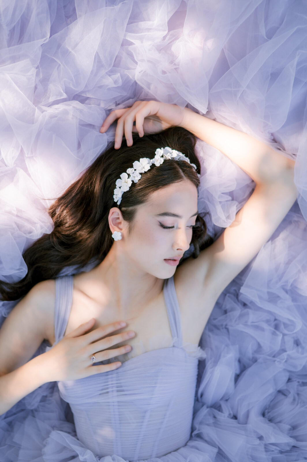 Model wearing a lavender tulle gown from Chic Bridals lies among soft ruffled fabric, wearing a white floral headband with eyes closed