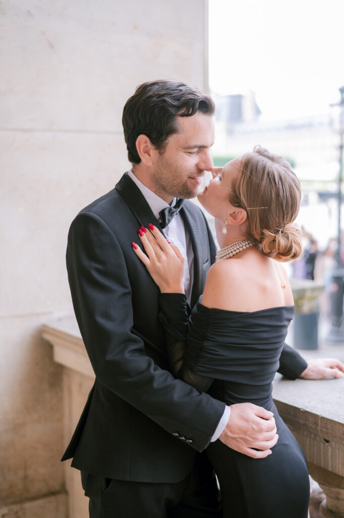 Couple in black formal attire embracing closely on a stone balcony, the man in a tuxedo and the woman in an off-shoulder dress, sharing an intimate moment.