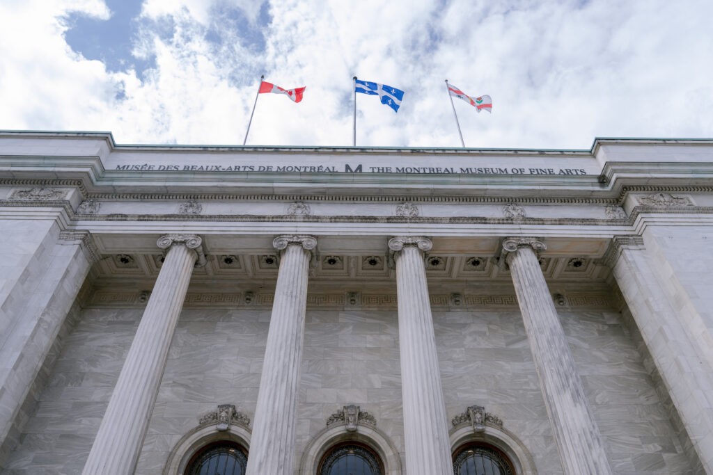 Low-angle view of the Montreal Museum of Fine Arts' neoclassical marble facade with grand columns, topped by Canadian, Quebec, and Montreal flags waving against a partly cloudy sky.