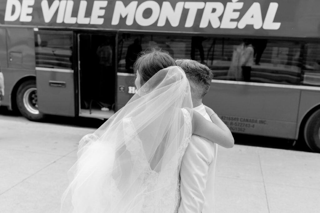 Black and white photo of a bride and groom from behind, her veil flowing in the wind, standing arm in arm on a Montreal sidewalk in front of a De Ville Montreal charter bus.