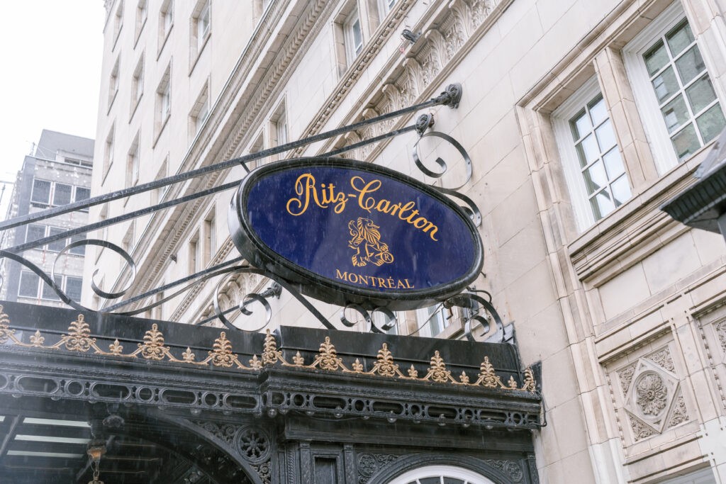 Ornate wrought-iron sign reading Ritz-Carlton Montréal in gold script on a navy oval background, mounted above the hotel's entrance canopy on a classic stone building facade.